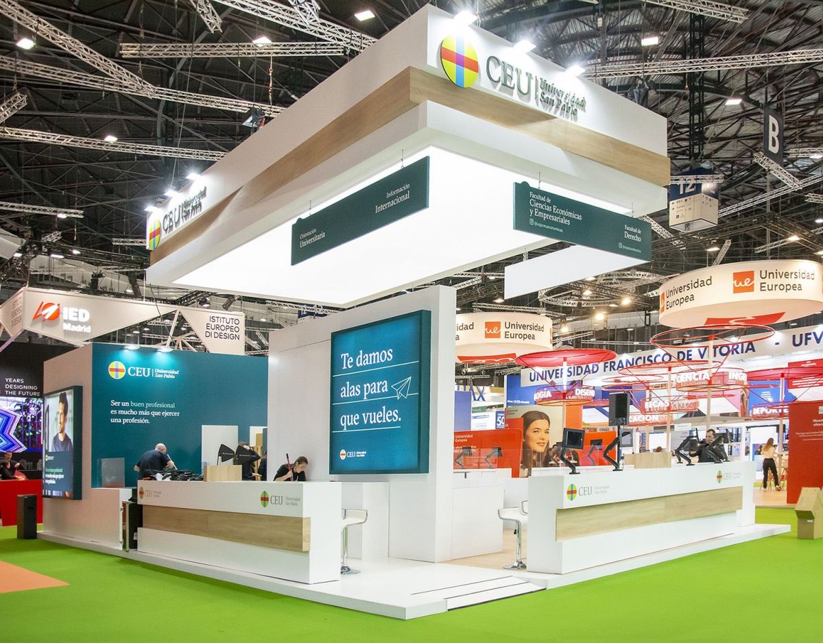 University exhibition stand in Madrid with 150m² island layout, perimeter counters, digital screens, and aerial lighting structure